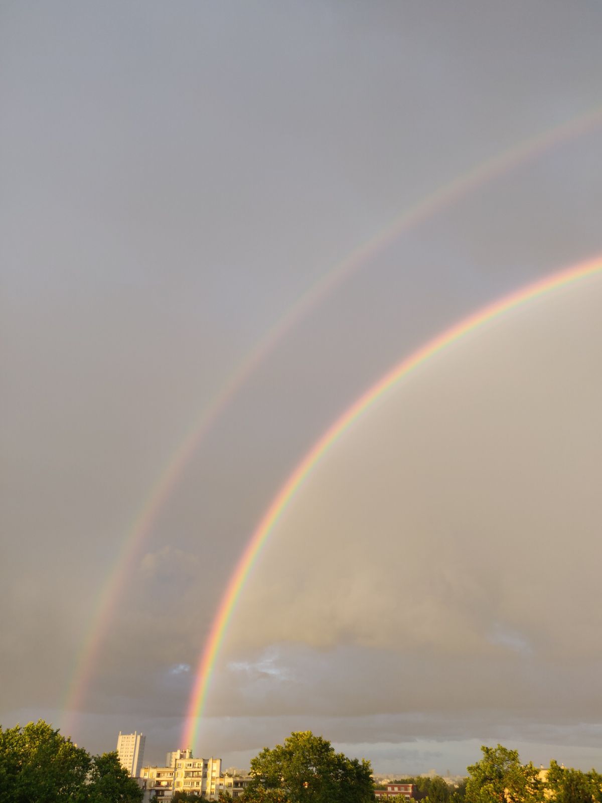 Double rainbow spanning above a city skyline under dramatic cloudy sky, showing the secondary arc clearly visible above the primary