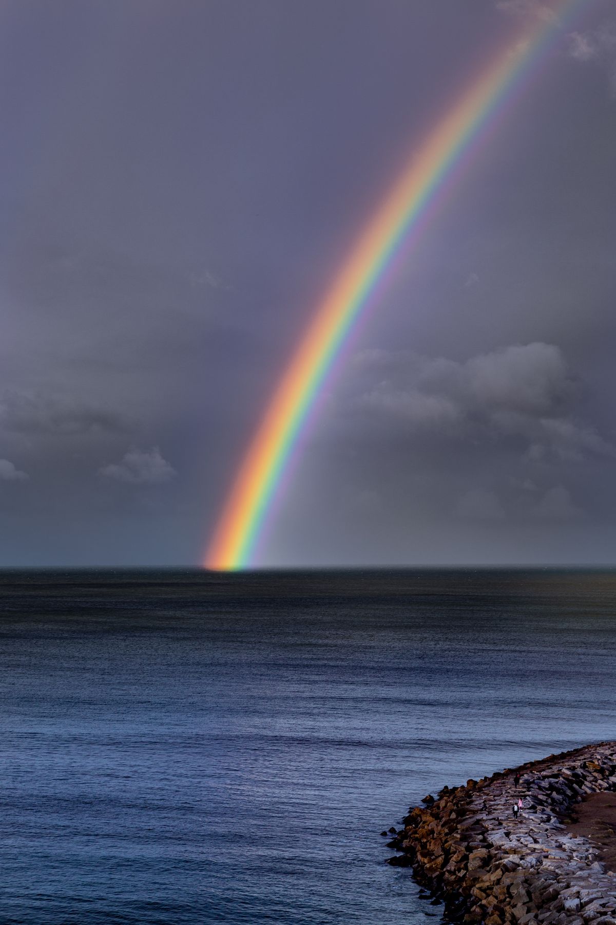 Stunning rainbow arches over ocean with rocky shoreline, showing the colours of the rainbow in order against a dramatic sky