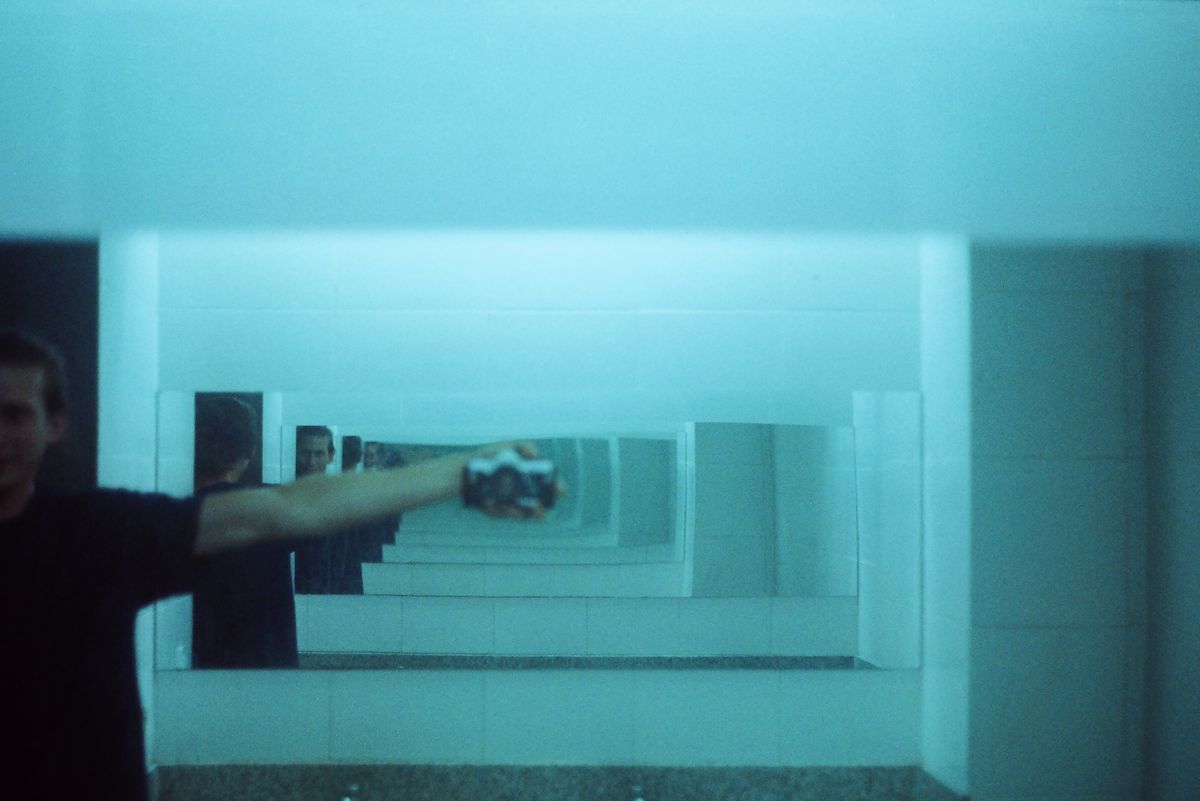 A man capturing his reflection in a bathroom mirror, creating multiple reflections that illustrate how plane mirrors produce virtual images at the same distance behind the mirror