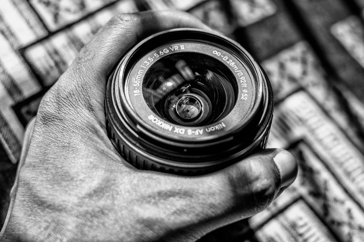Black and white image of a Nikon camera lens being held, showing the convex lens at the front