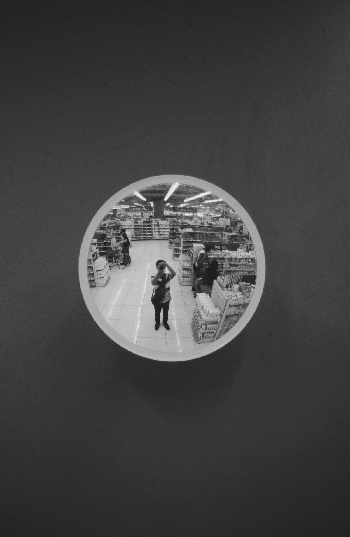 Black and white photograph of a photographer taking a picture reflected in a round security mirror in a supermarket, demonstrating how convex mirrors are used for surveillance