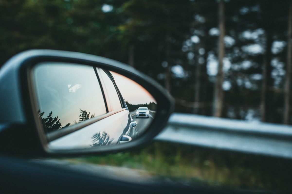 Close-up shot of a car side mirror on a scenic forest road, showing the type of convex mirror used in vehicles to give a wide-angle view